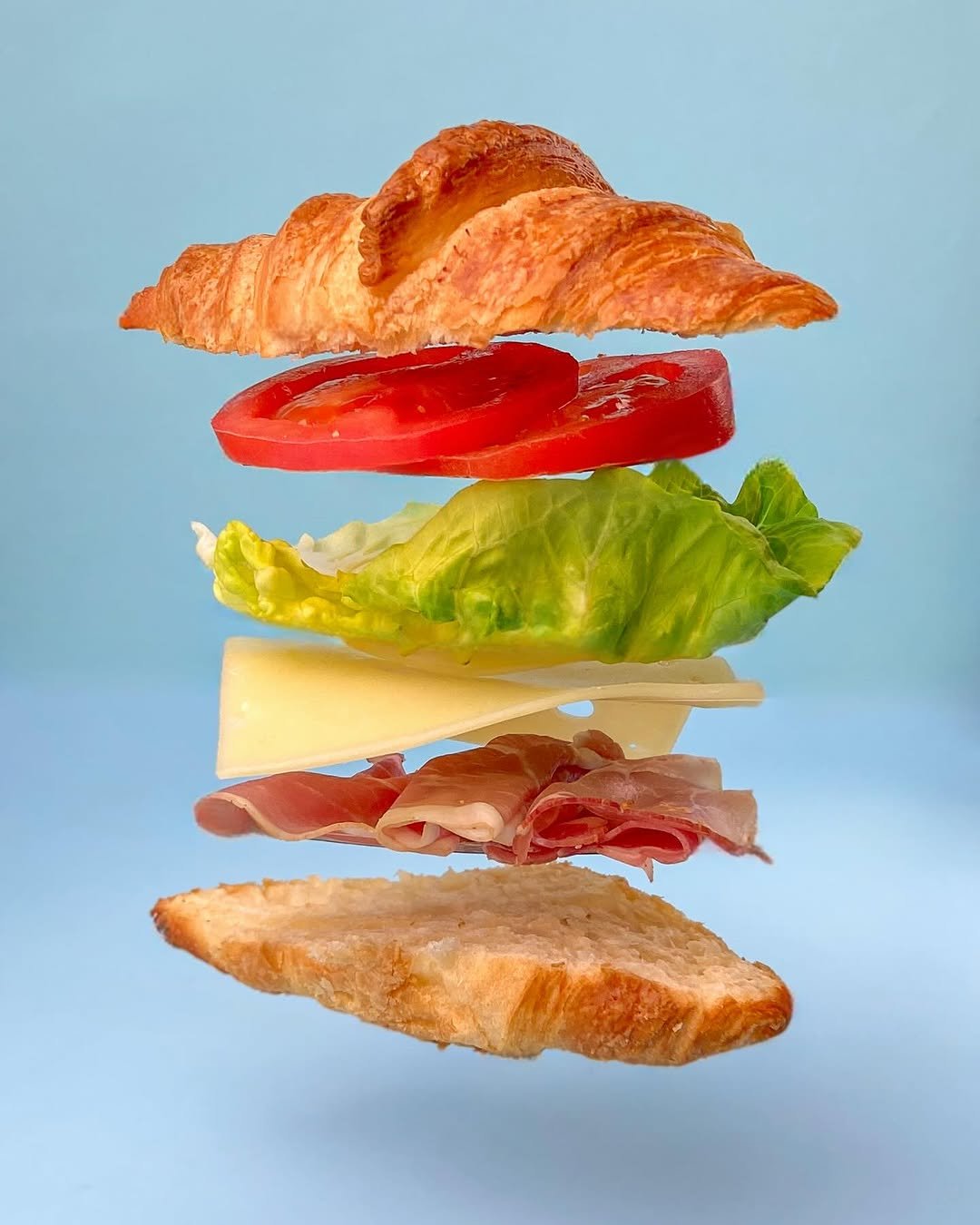 The ingredients of a sandwich are suspended in mid-air on fishing line with a sky blue background. There is a croissant, tomato, lettuce, cheese, and prosciutto.