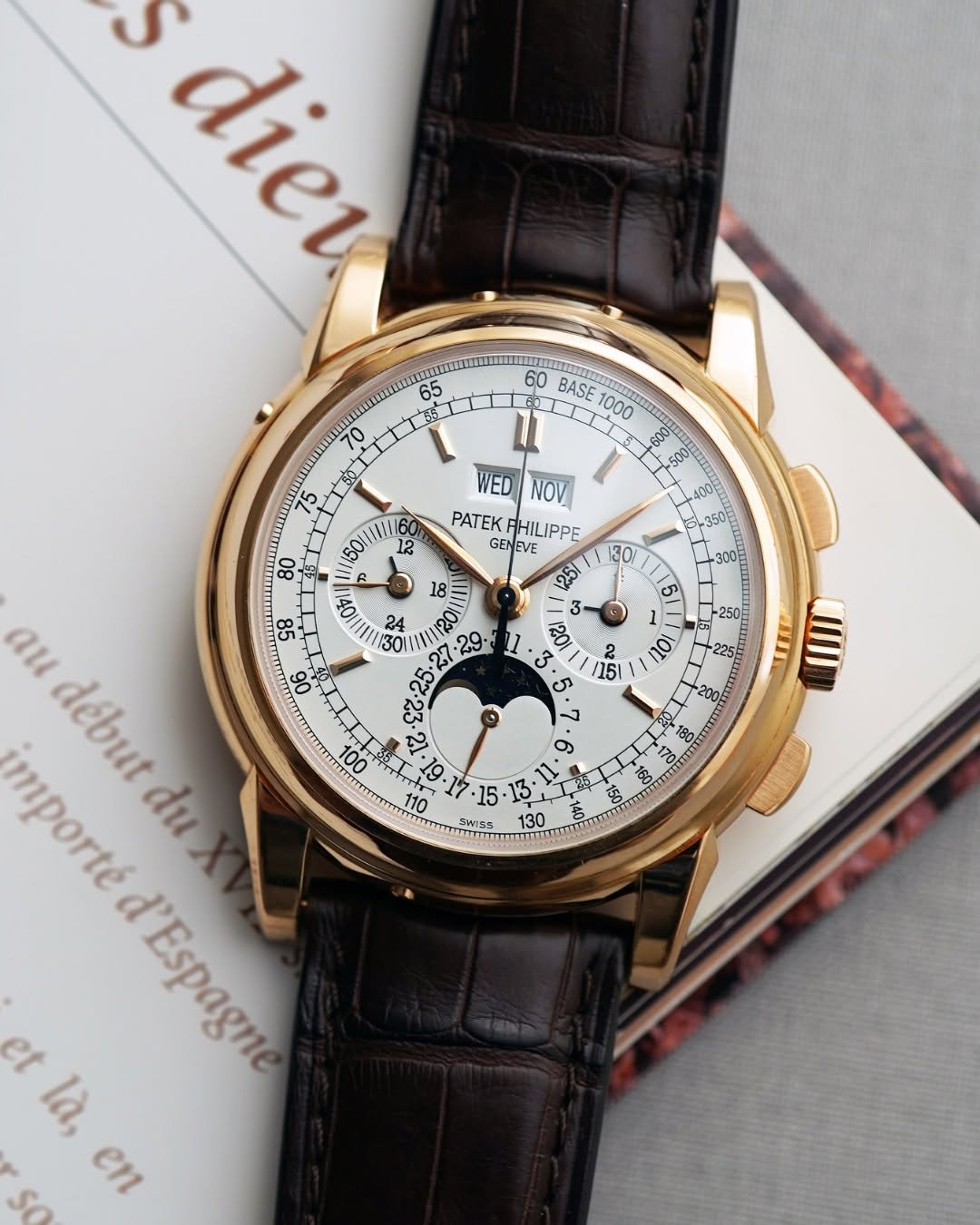 Chrono24 luxury watch with a gold case and brown leather strap, displayed on an open book. The watch face features multiple dials, including day and month indicators, a moon phase display, and intricate markings. The design is classic and refined.