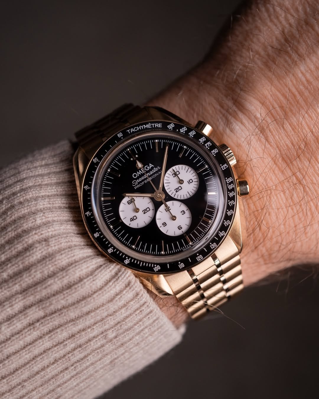 Chrono24 a gold Omega Speedmaster Professional watch is on a wrist. It features a black dial with three white subdials and a tachymeter scale on the bezel. The watch is paired with a gold bracelet, and the wearer has a light-colored, ribbed sweater.