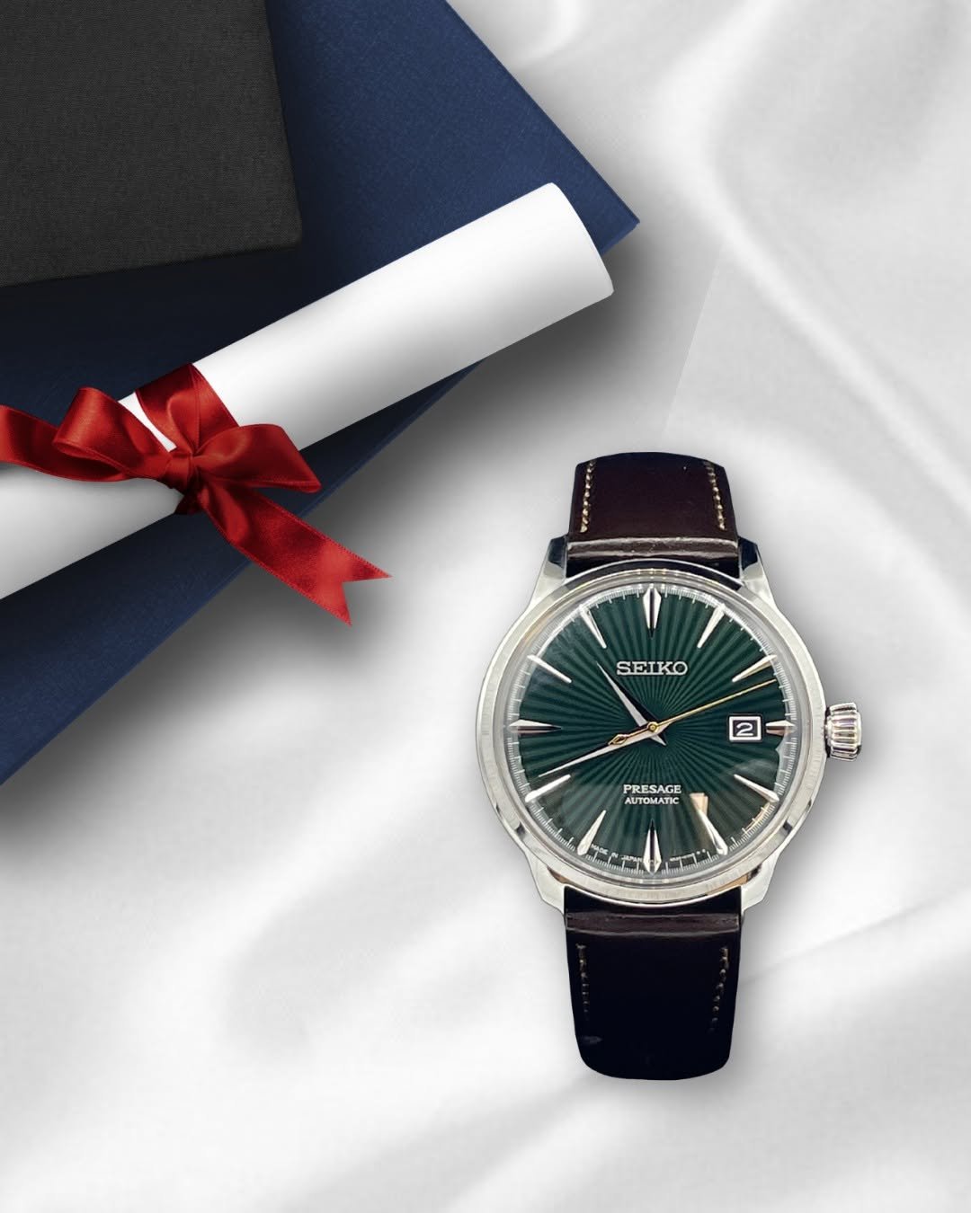 Chrono24 a Seiko Presage Automatic watch with a green dial and dark leather strap is placed on a white satin fabric. Beside it, a rolled diploma tied with a red ribbon and a stack of blue and black fabric-covered books are visible.