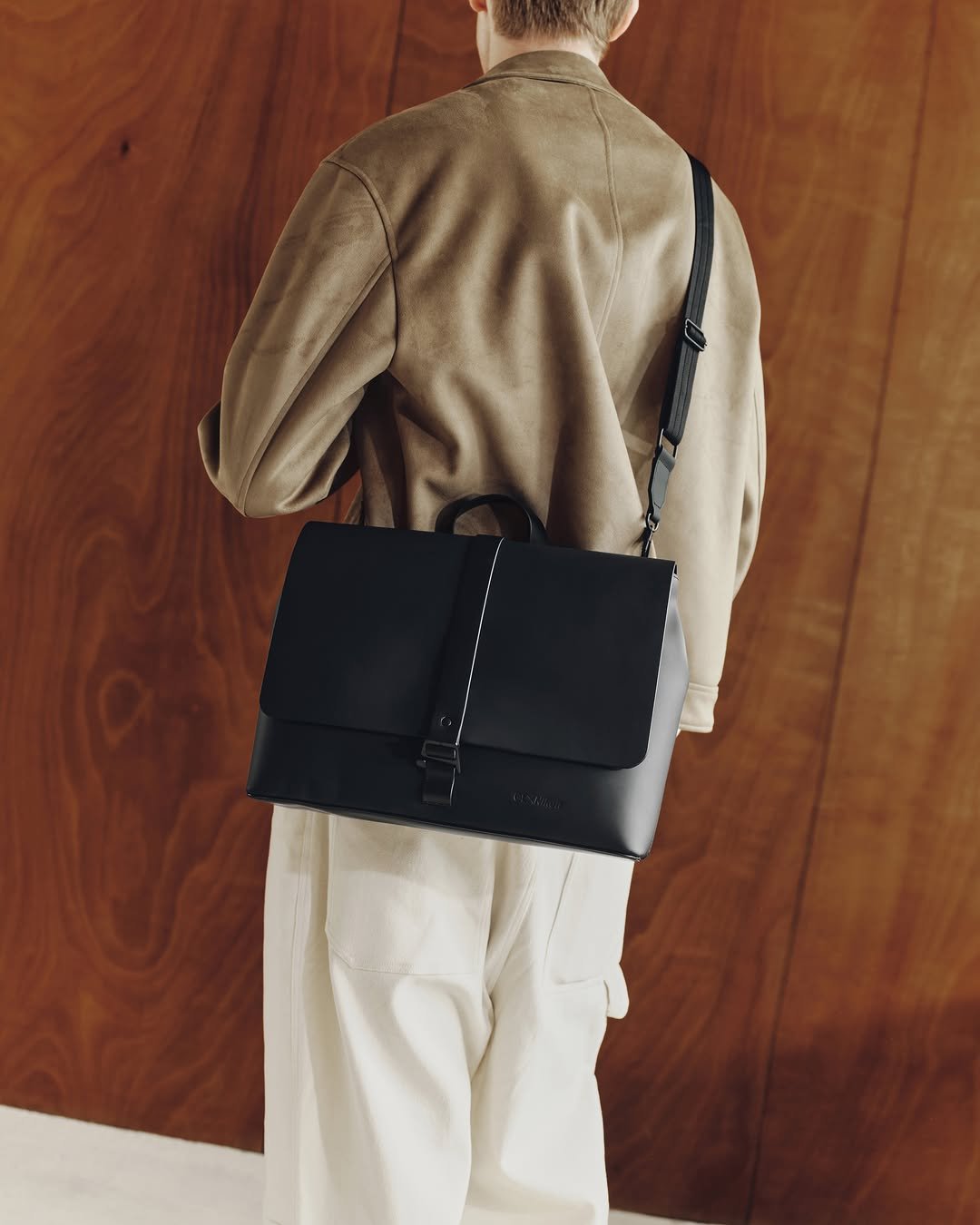 A person wearing a tan suede jacket and white trousers seen from behind, carrying the black GL X Nikon Messenger Bag over their right shoulder, against a warm wood-paneled background.