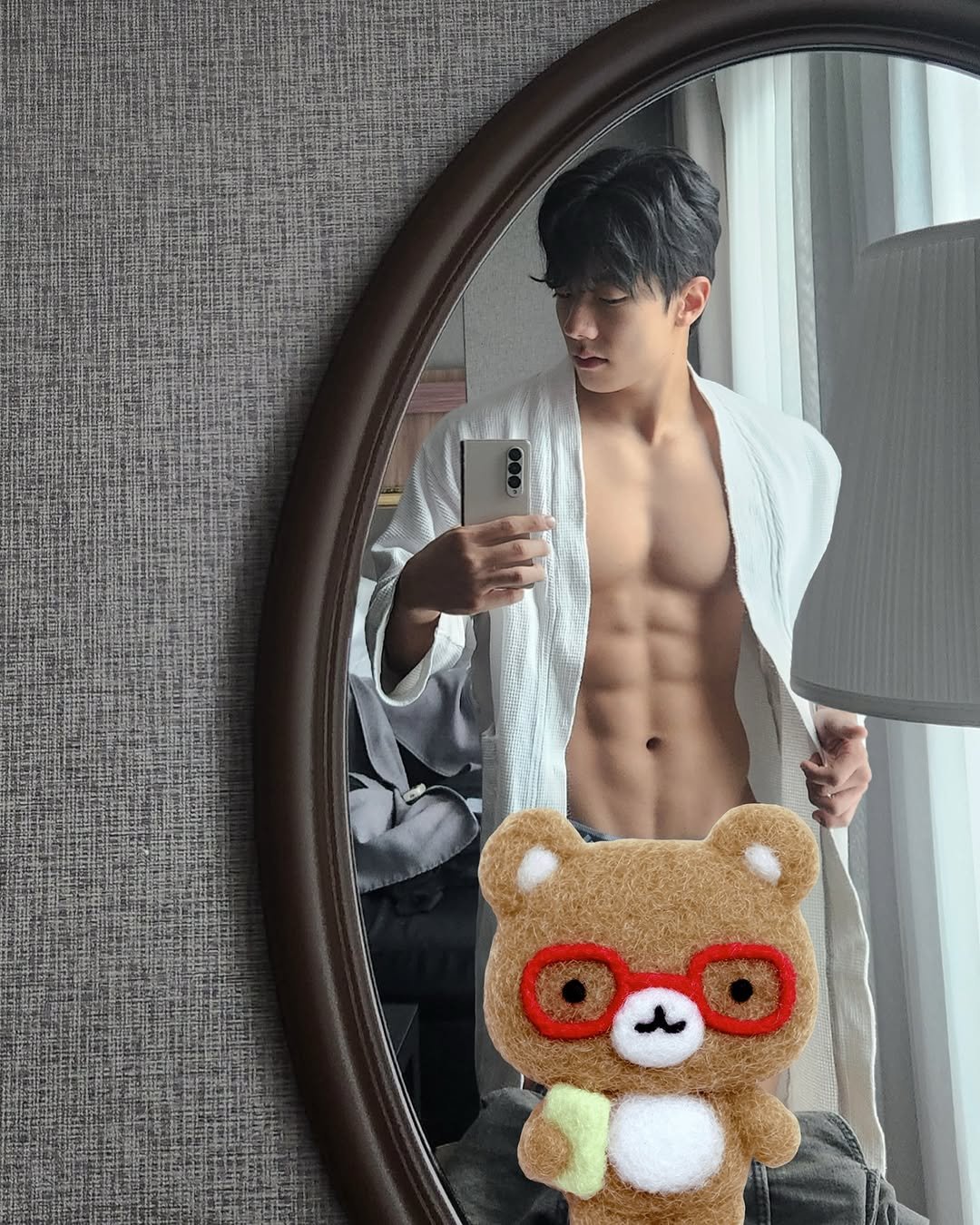 A mirror selfie of a bodybuilder highlighting abs and upper body muscles after workout. A cute bear character sticker with red glasses is placed in the front, making the photo look friendly and playful.