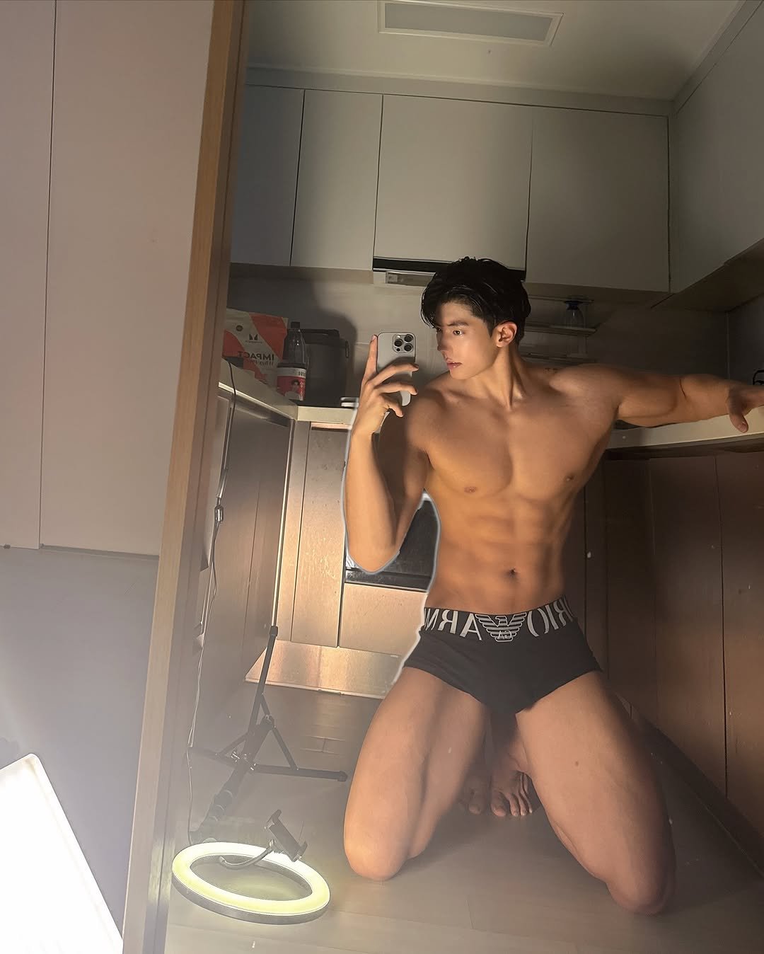 A male bodybuilder kneeling on the floor in front of a mirror, wearing black boxer briefs, showing defined muscles and athletic physique in a fitness-themed self-portrait.