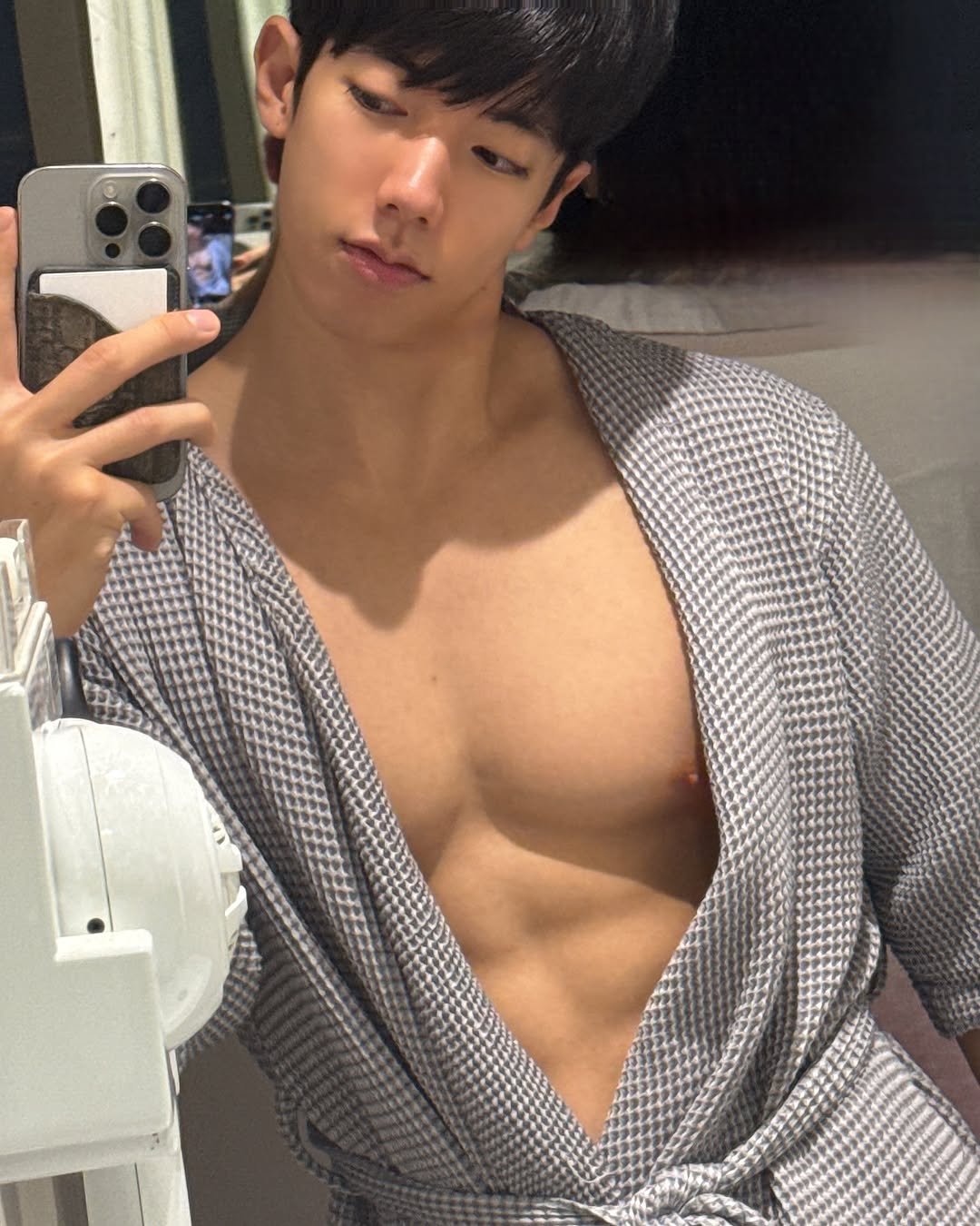 A professional bodybuilder taking a mirror selfie in a gym-style pose, wearing a gray robe and showing a strong, athletic upper body.