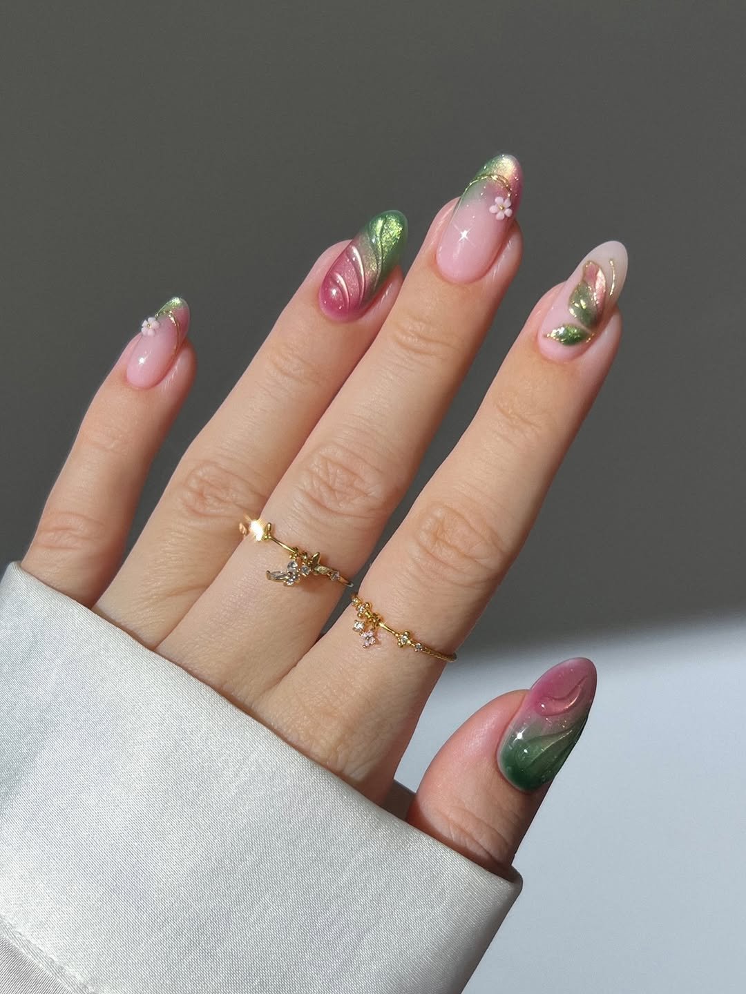 Pink and green glass fairy nails, butterfly nails, spring nails, flower nails, ombre gradient nails, gold nails, french tip nails, milky pink nails, dainty gold ring with dangling butterfly and flowers, beige pink cat eye nails with gold shimmer and white 3D texture flowers, gold open leaf ring with gems, scattered diamond eternity gold ring, pastel green and pink aura nails, 3D floral nails, gold pearl ring, curved wedding band with diamonds,