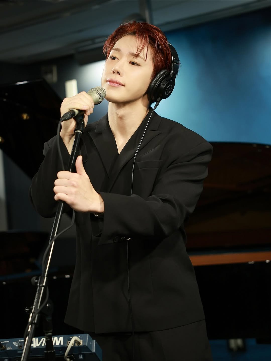Wearing headphones, Wonho stands at a microphone with a piano in the background.