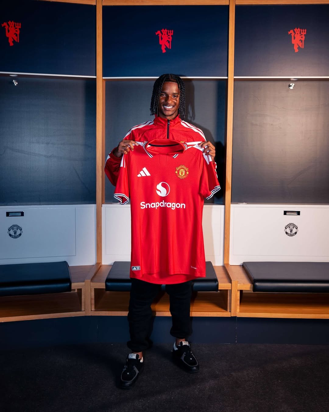 Tynan Thompson grins while holding our red 2026/27 Manchester United home shirt inside the dressing room with club logos visible.