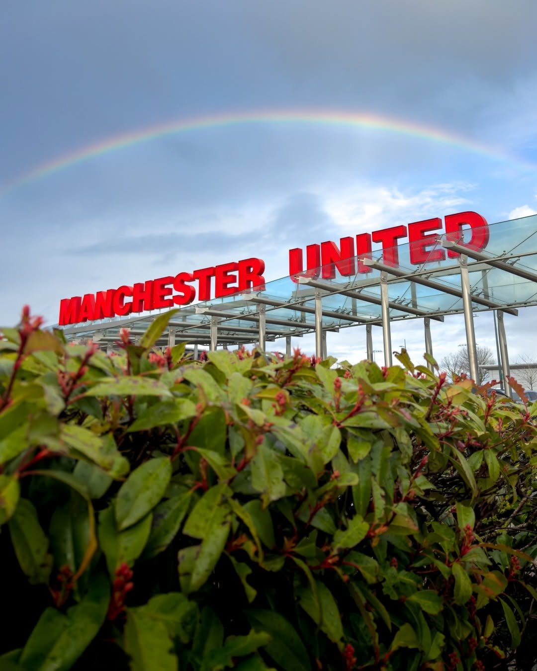 Red Manchester United sign atop a stadium entrance with a rainbow arching across a cloudy sky.