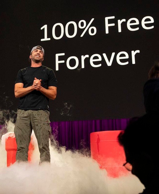 Mark Rober stands on stage with fog effects, wearing casual clothes and a backward cap. Behind him, a large screen displays the words "100% Free Forever." Red props are visible, and the setting suggests it is at TED