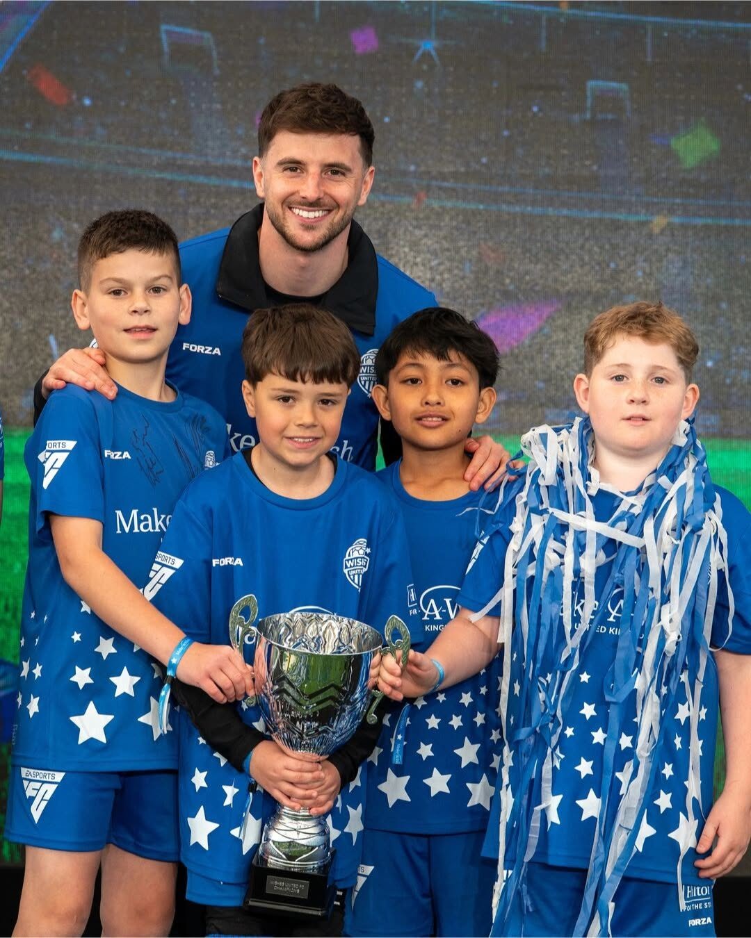 A group of wish children in blue star-patterned Wishes United FC kits stand on a stage holding a trophy. England and Manchester United footballer Mason Mount stands behind them, smiling. The background shows a stadium scene and there is confetti.