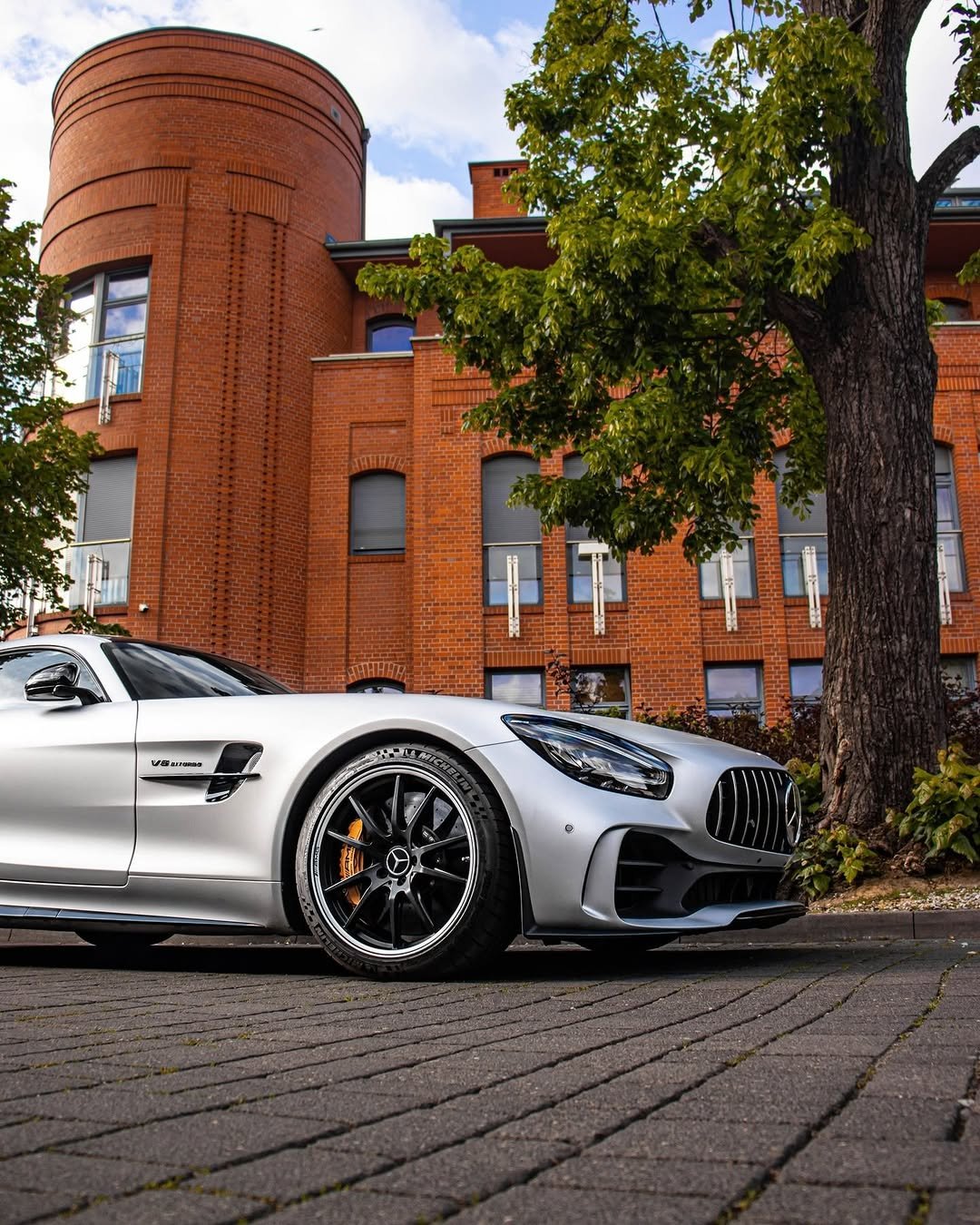 A silver sports car is parked on a cobblestone street. In the background, a red brick building with large windows and a rounded structure can be seen, alongside a green tree.