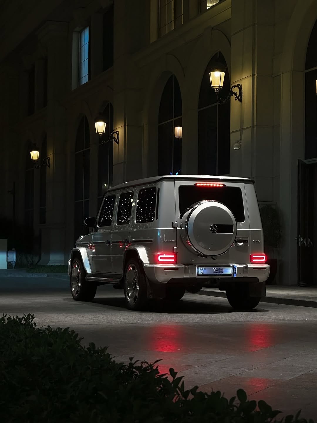 At night, a silver G-Class with a spare tire mounted on the back stands before a building lit by ornate lamps. The vehicle's taillights glow red, and its license plate reads '7918'.