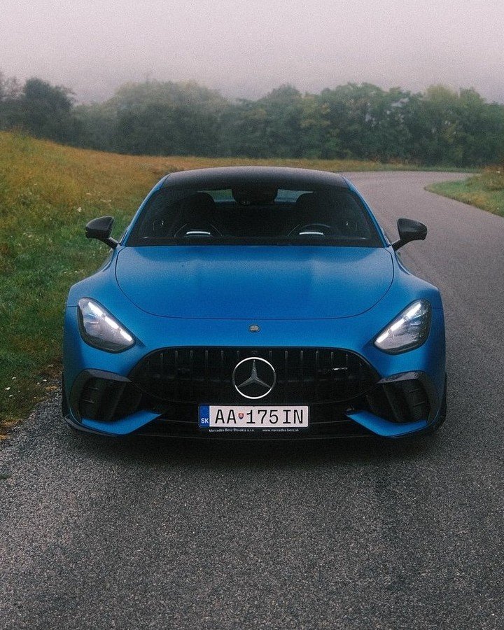 A blue Mercedes-AMG GT is parked on a winding road surrounded by lush green grass and trees, under a foggy sky. The car faces forward, showcasing its sleek design and prominent front grille.