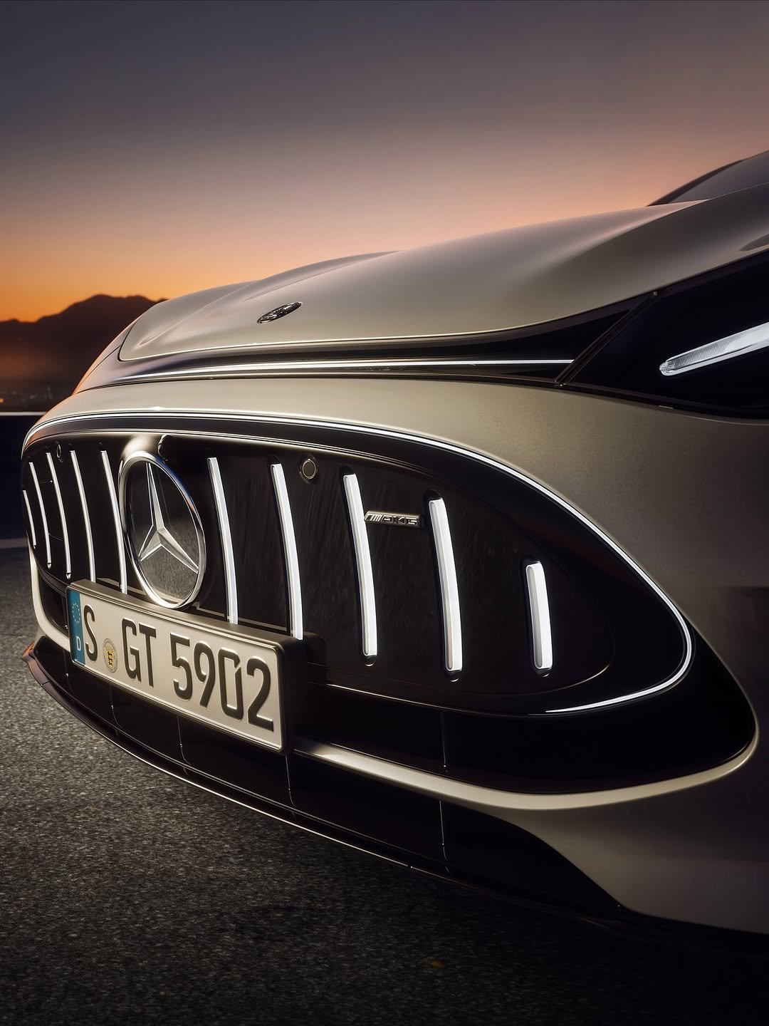 Close-up of the front of a silver Mercedes-AMG car with an illuminated grille featuring a large Mercedes logo and an AMG badge. Mountains and city lights silhouette against an orange-purple sunset sky.