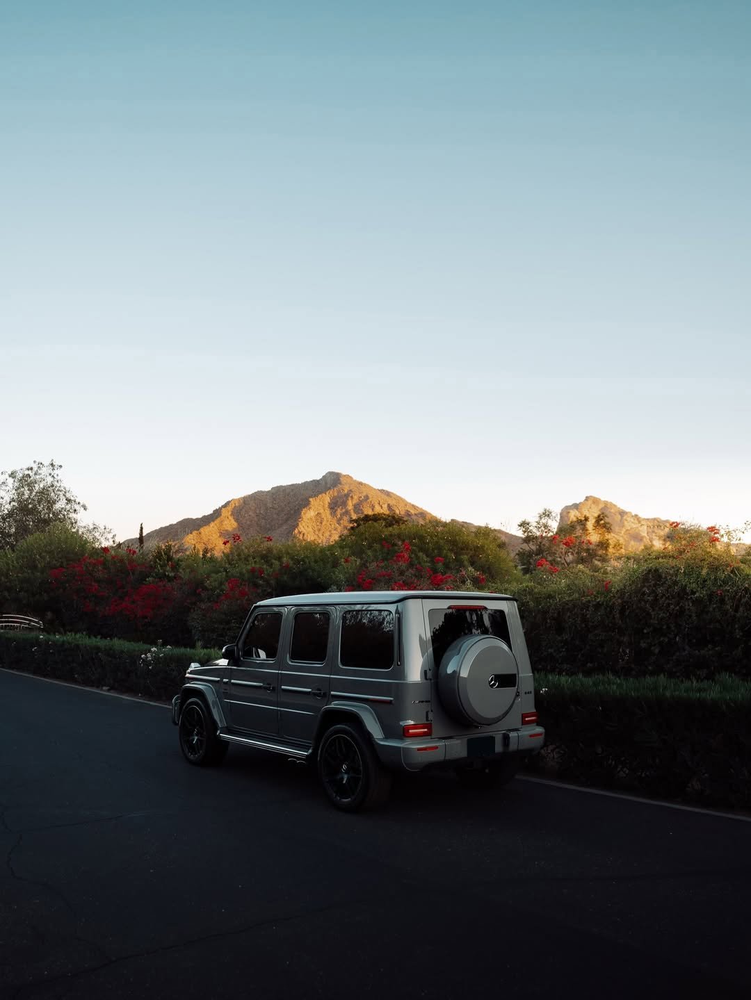 A silver Mercedes-Benz G-Wagon SUV is parked on a dark asphalt road, seen from the rear. Green hedges with red flowers line the road. In the distance, sunlit mountains rise under a clear blue sky.