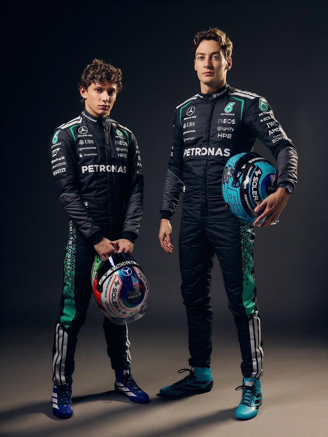 Kimi Antonelli and George Russell stand side by side wearing our 2026 black race suits. Kimi and George are holding their race helmets