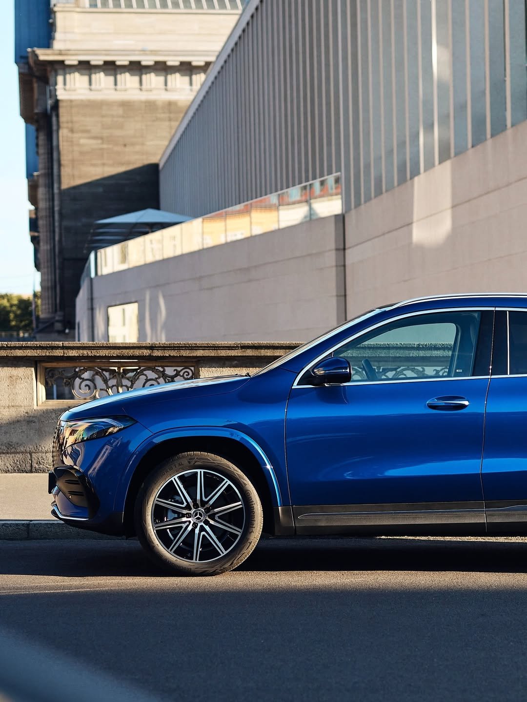 A sleek, deep blue SUV is parked on a city street, brightly lit by sunlight. The car's glossy surface reflects the blend of modern and traditional buildings in the background. A Mercedes-Benz logo is visible on the front wheel, with tire details 'Continental EcoContact 6' and '235/50 R 19 T XL'.