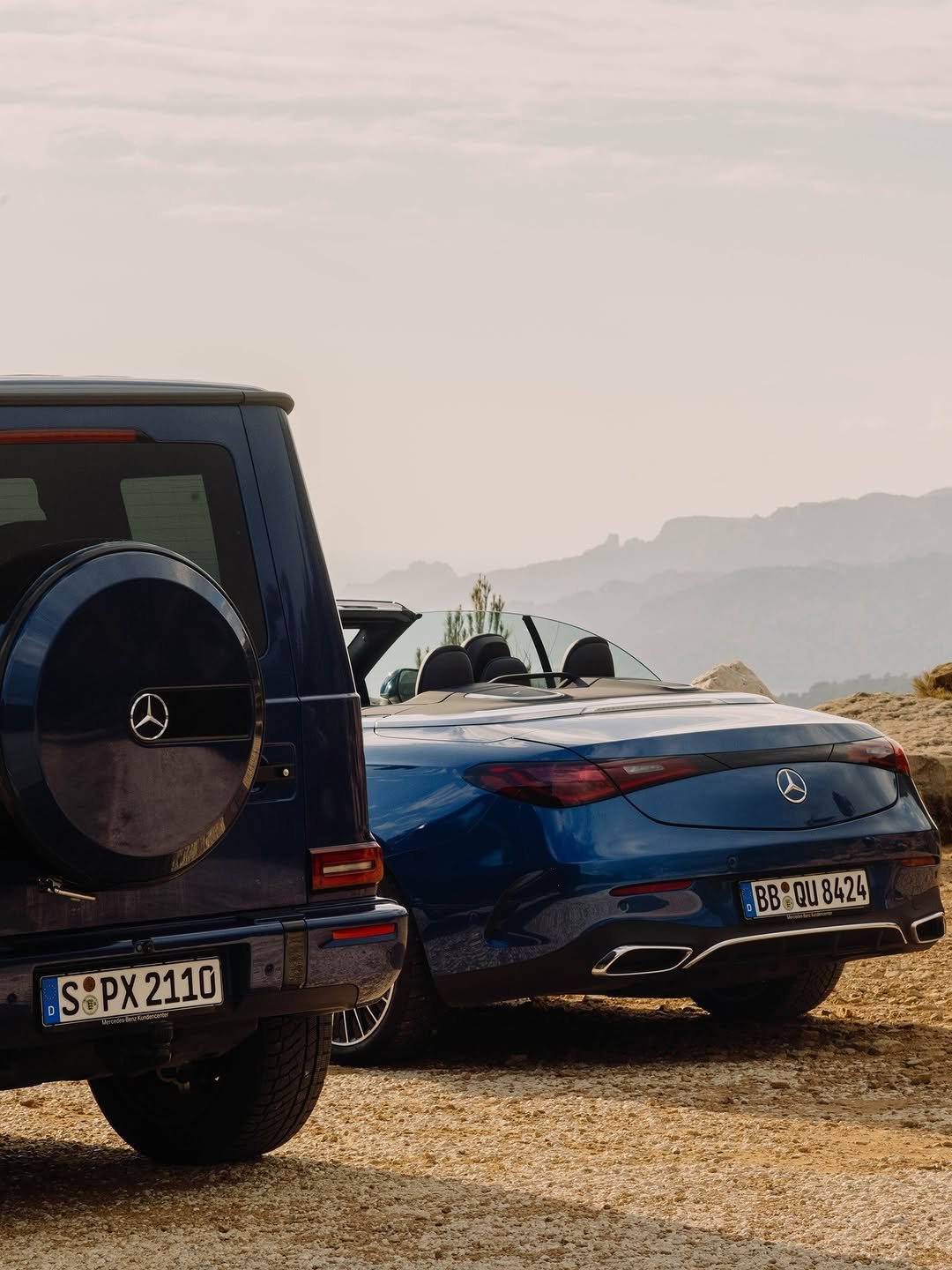 Two blue Mercedes cars, a G-Wagon and a convertible parked on a dirt surface against a backdrop of hazy mountains.