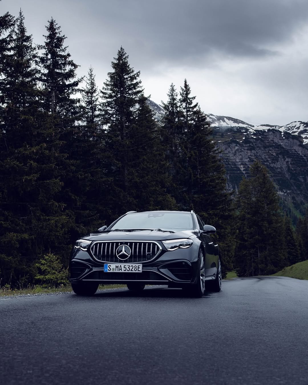 A black Mercedes-Benz E-Class is parked on a winding road surrounded by tall pine trees. Snow-capped mountains rise in the background under a cloudy sky.