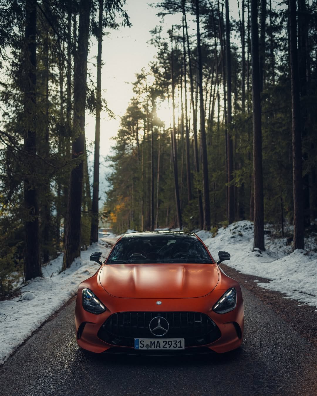 A sleek orange Mercedes sports car parked on a snow-covered road, surrounded by tall evergreen trees. The scene is illuminated by soft, diffused light from the setting sun in the background.