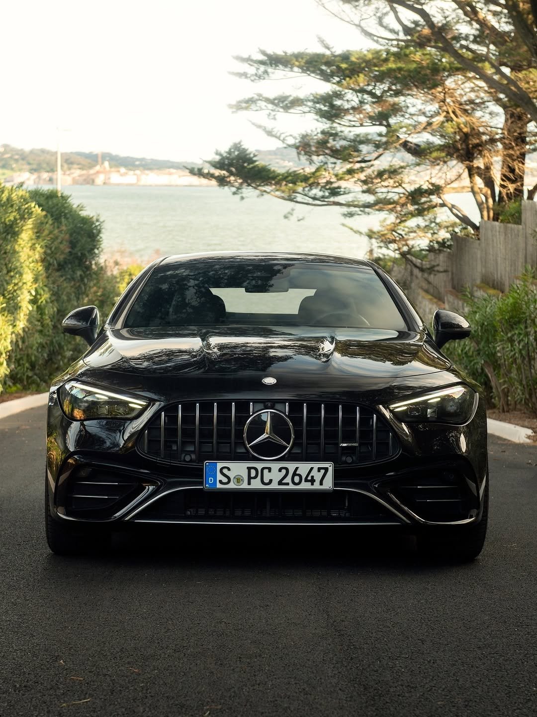 Front view of a glossy black Mercedes-Benz AMG car on an asphalt road. It has LED headlights, a vertical grille with a large Mercedes star, and license plate "S PC 2647". Lush greenery frames a background of water, distant city buildings, and a bright sky.