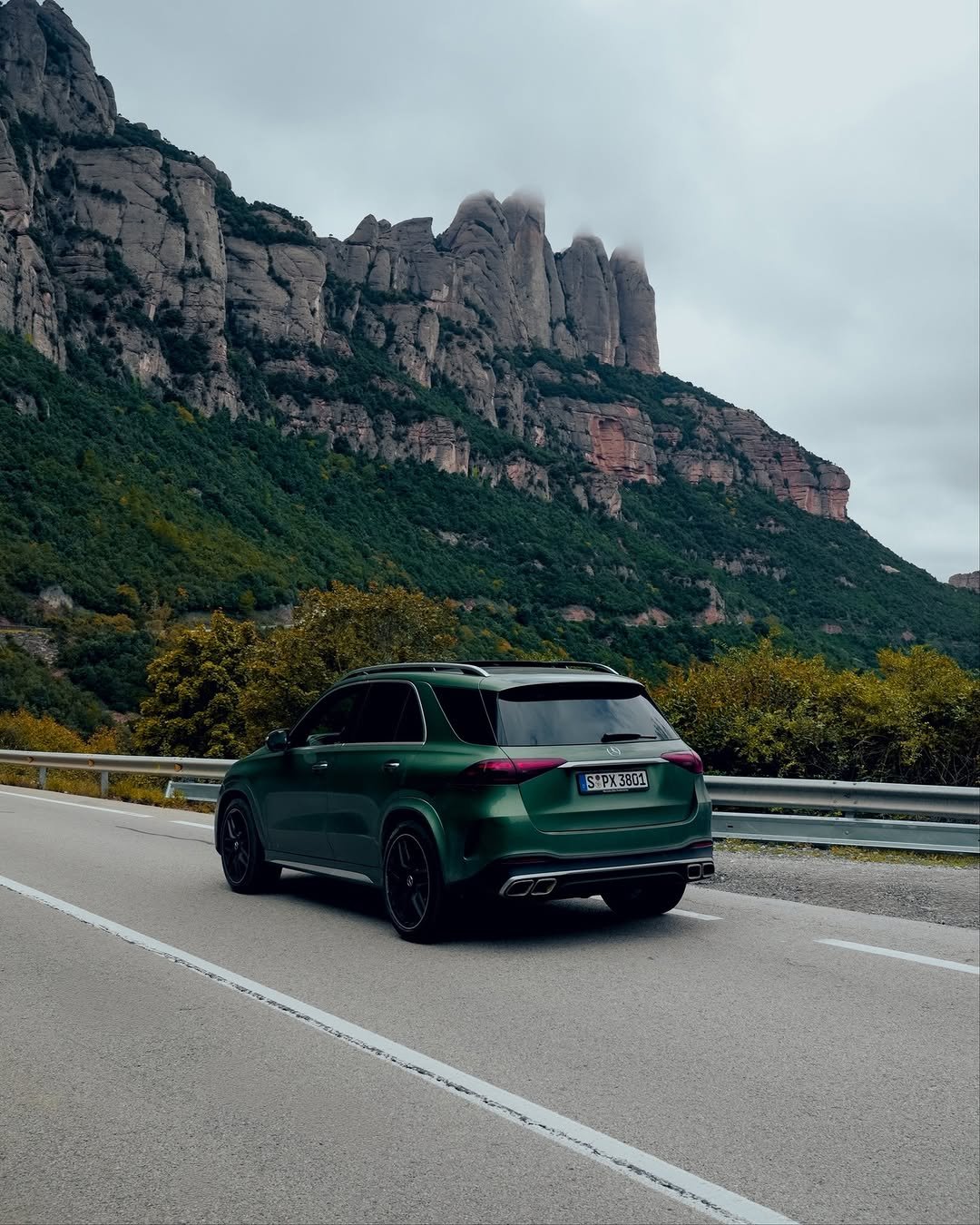 A green GLE parked on the side of a winding road, with tall rocky cliffs and lush green trees in the background. The sky is cloudy, suggesting an overcast day. The vehicle's rear shows sleek lines and dark wheels, emphasizing its sporty design.