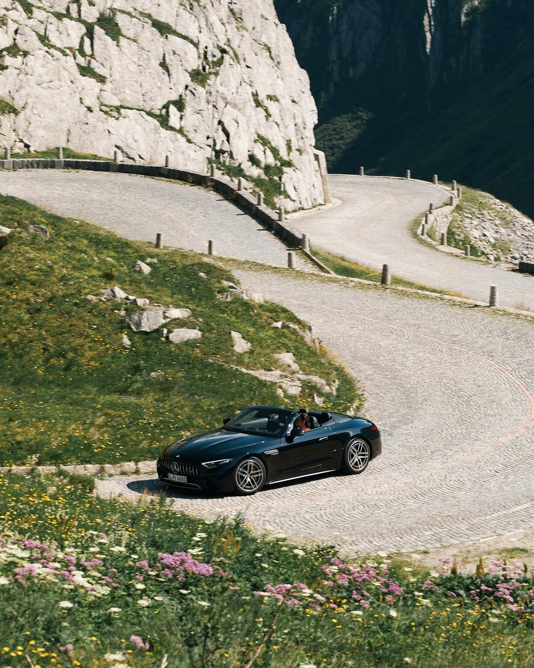 A black convertible Mercedes-Benz, license S PV 4265, drives on a winding cobblestone mountain road. The road features hairpin turns and is flanked by green grass with yellow and purple wildflowers, and imposing rocky cliffs. A person is driving the car.