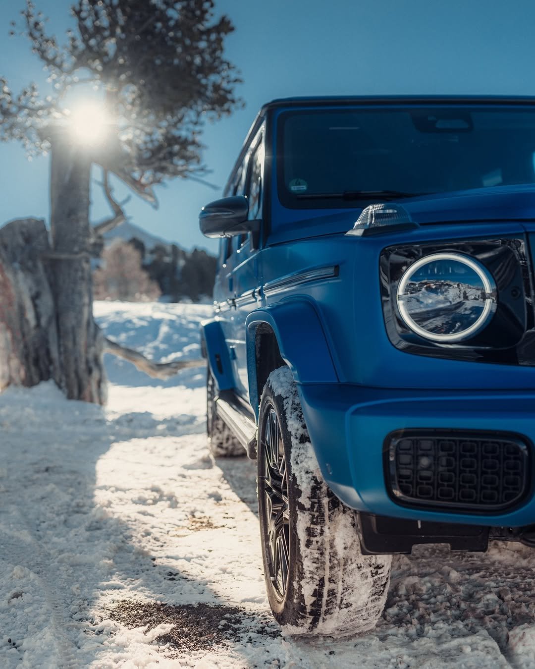 A blue electric G-Class is parked on a snowy path, partially obscured by snow. The sun shines brightly in the background, illuminating the area. Trees are visible nearby, and the SUV's wheels show traces of snow.