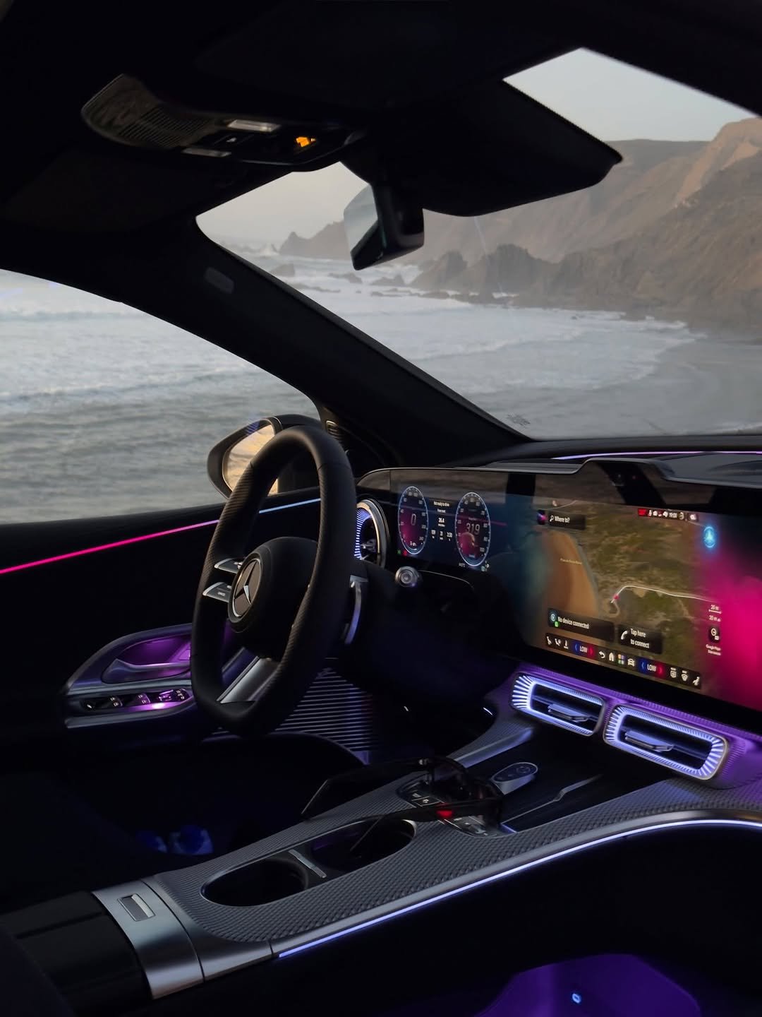 Interior of a car with glowing accents, dashboard displaying a map with text "Where to?", speedometer showing "0" and "319", indicating speed. "No device connected" text is also visible. The car is parked overlooking the sea.