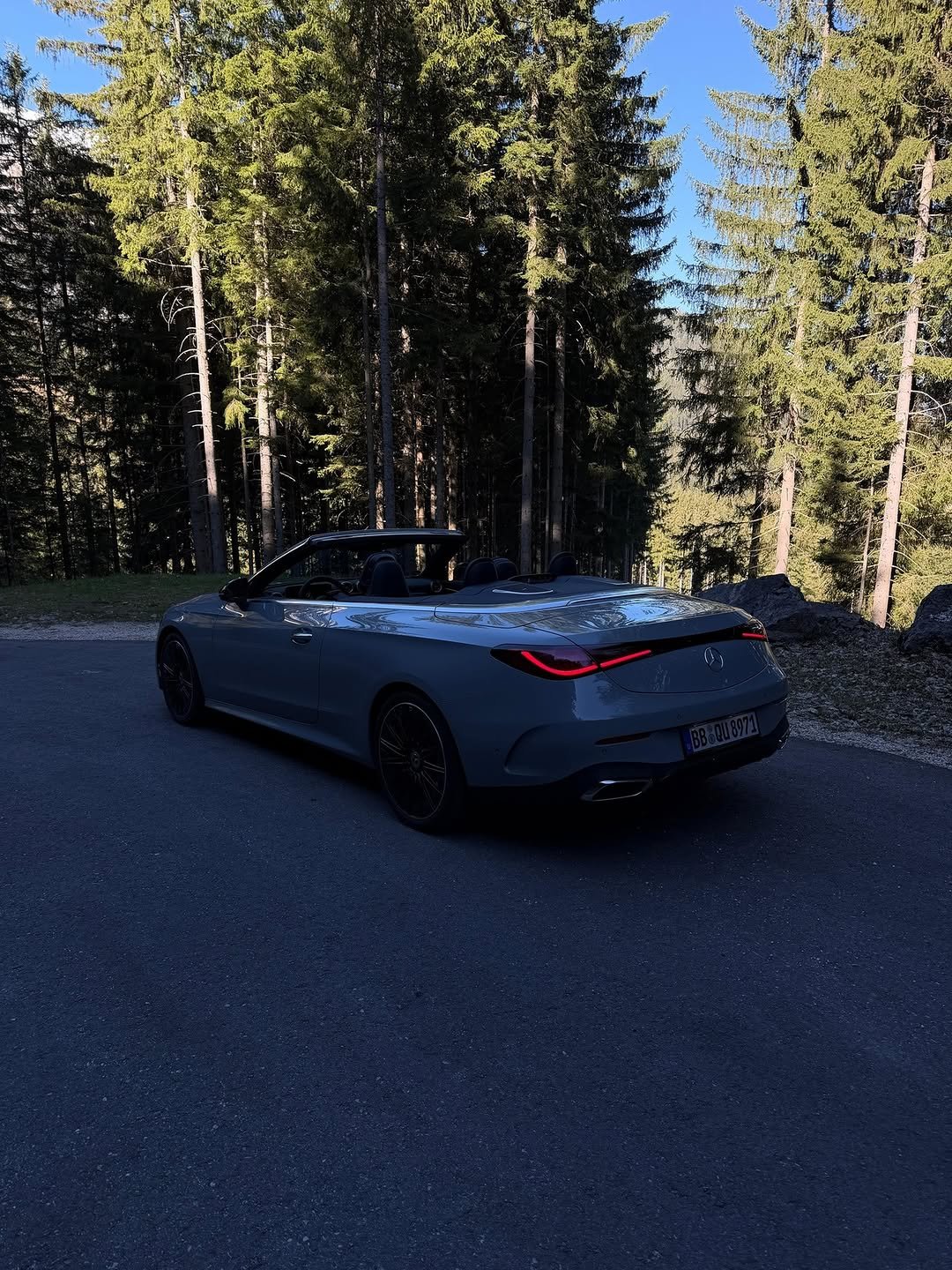 A light blue Mercedes-Benz convertible with its top down is parked on a dark asphalt road. The car faces away from the viewer, its red taillights glowing. Dense, sunlit pine trees line the background.