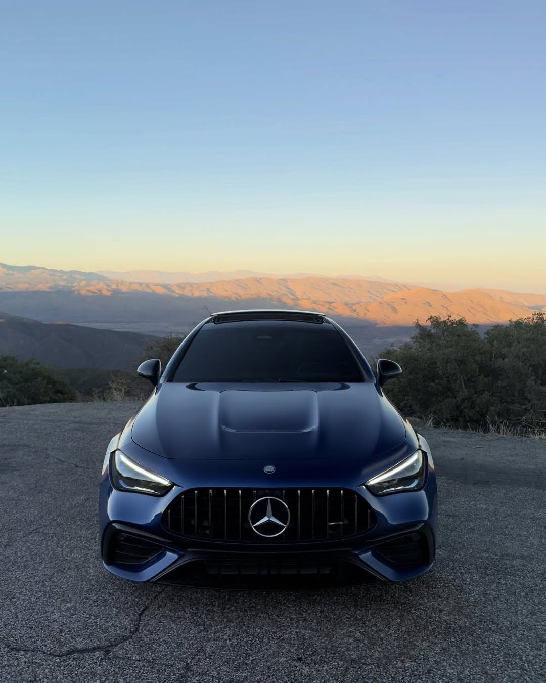 A blue Mercedes-Benz car is parked on a scenic overlook. The vehicle faces the camera with a mountainous landscape in the background, showcasing golden hues from the setting sun. The sky is clear, adding to the serene atmosphere.