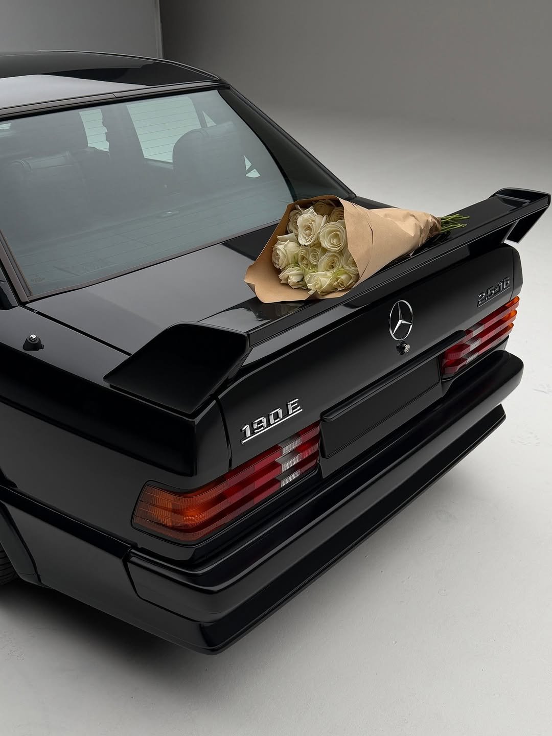 Rear view of a black Mercedes-Benz 190 E on a white seamless background. A bouquet of white roses wrapped in brown paper rests on the trunk.