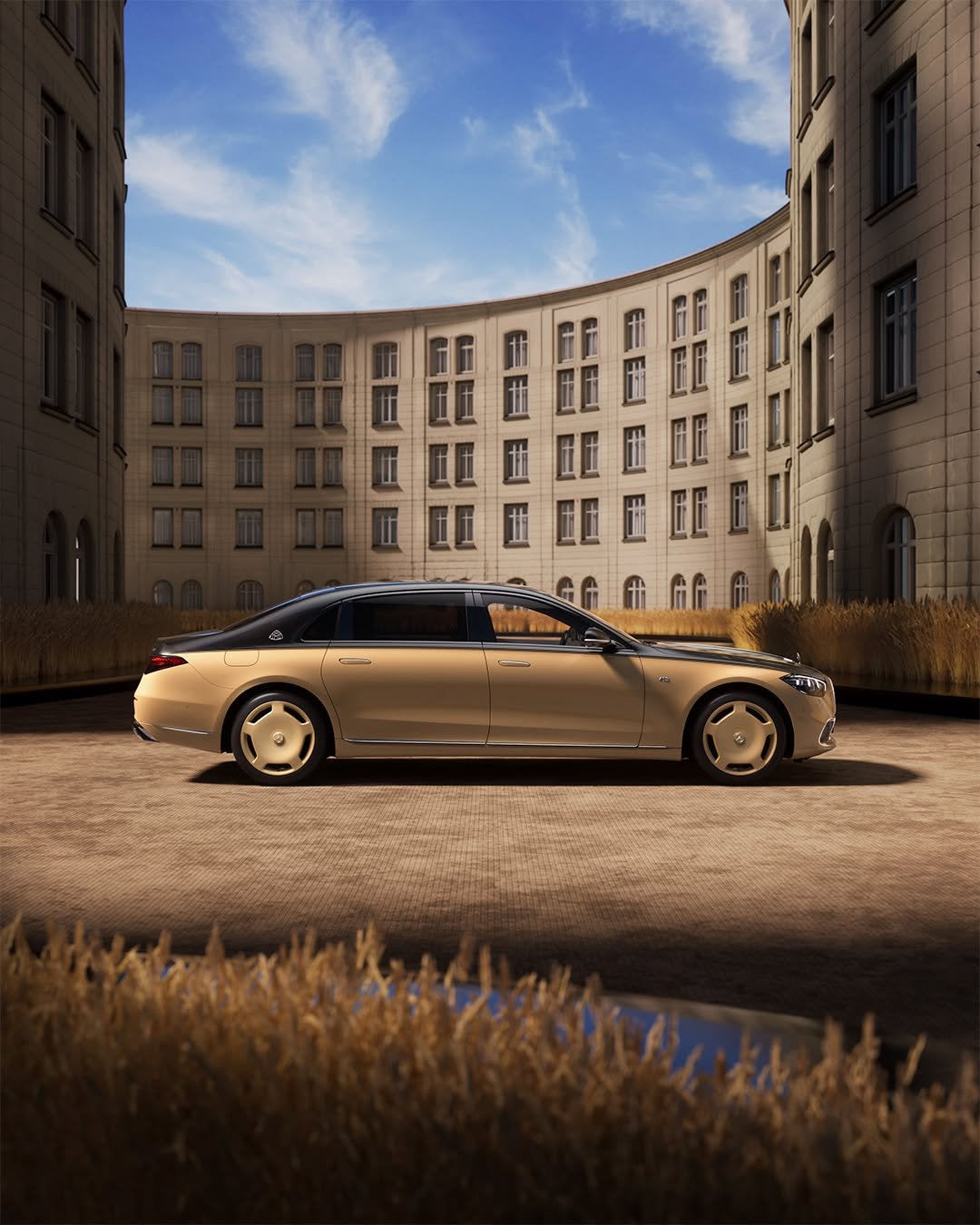 The Mercedes-Maybach S-Class is parked in a courtyard surrounded by tall, modern buildings. The car is golden with sleek lines, reflecting the bright blue sky with scattered clouds. Tall grasses and a clear surface in the foreground add to the serene atmosphere.