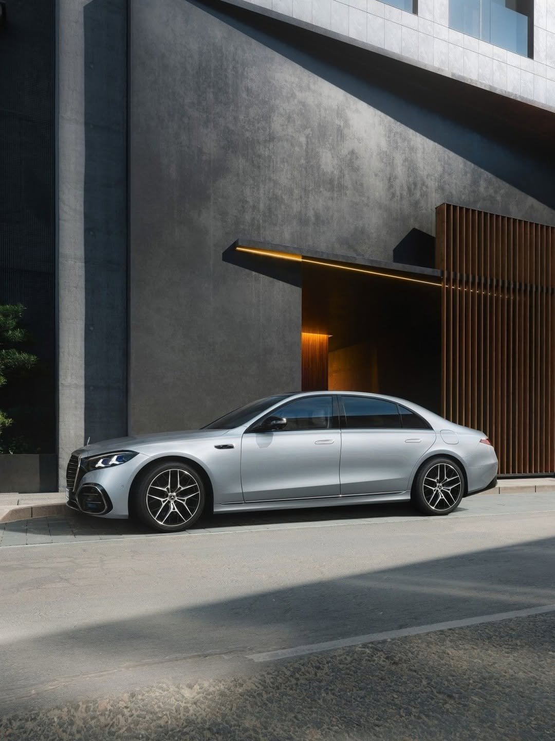 A sleek silver Mercedes-Benz S-Class sedan is parked curbside next to a modern building. The building features dark grey textured walls, an illuminated orange light strip above its entrance, and vertical wooden slats. Sunlight creates strong shadows.
