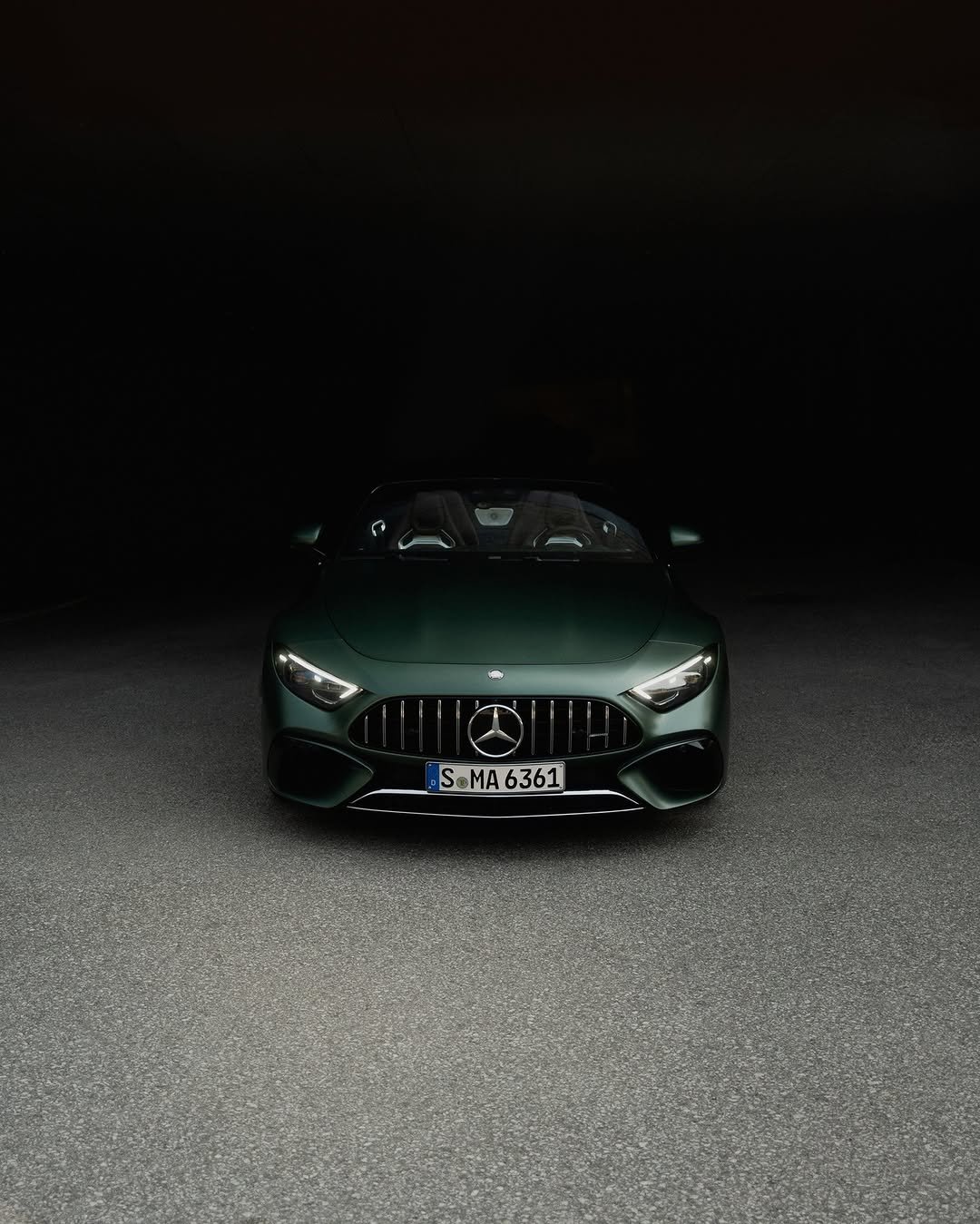 Dark green sports car with headlights on, facing forward in a dimly lit tunnel. The top is down, revealing the interior. License plate: S MA 6361.