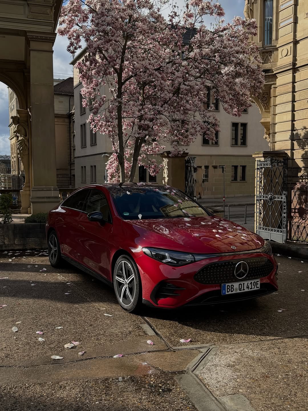 A red Mercedes-Benz car, possibly an EQE, is parked under a blooming magnolia tree with pink flowers. Petals are scattered on its hood, windshield, and the paved ground. Historic buildings with arched entrances and an ornate gate are in the sunlit background. License plate: BB QI 419E.