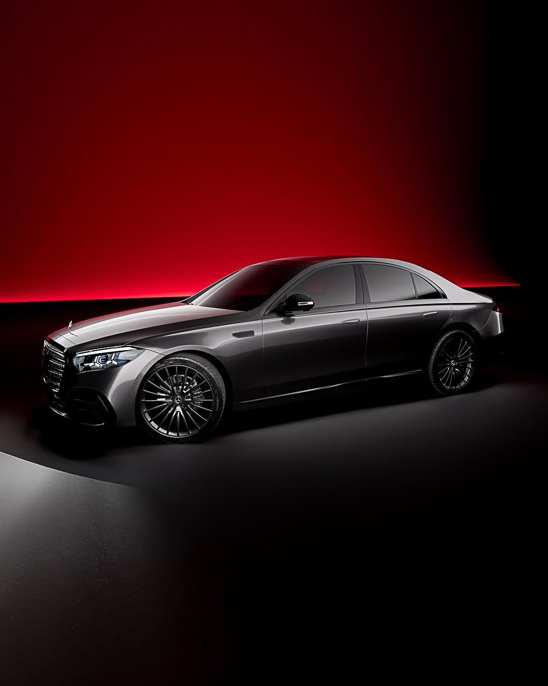 A sleek, dark grey sedan with active headlights and intricate black wheels sits on a dark floor. A dramatic red light illuminates the wall behind it, creating a sophisticated contrast.