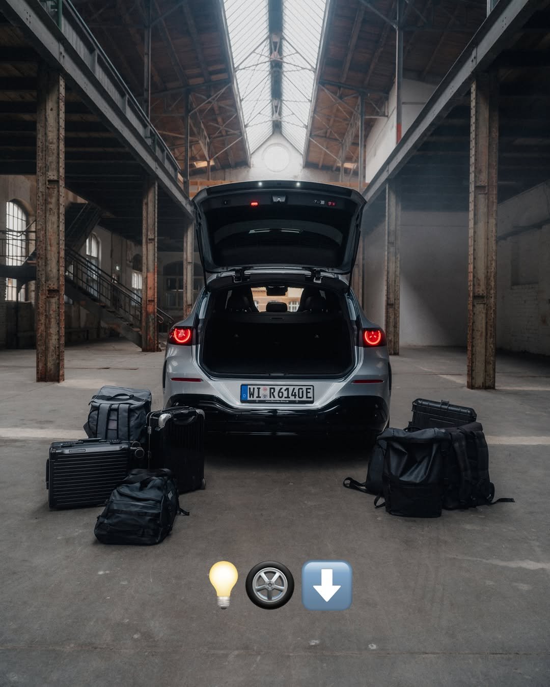 Silver car with open trunk packed with black luggage in a large, atmospheric industrial building. License plate reads WI R 6140 E. The building features high ceilings, skylights, and exposed metal beams.