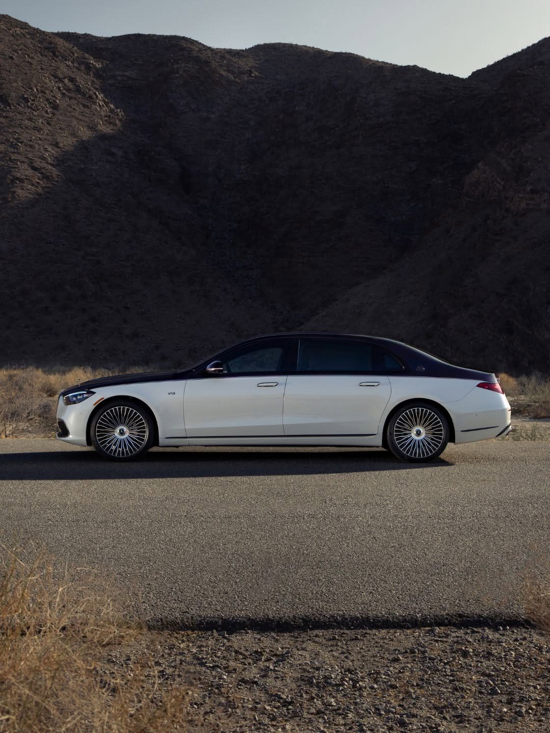 White and black Mercedes-Maybach V12 luxury sedan parked sideways on an asphalt road. Dark, rugged mountains loom in the background under a clear sky, with sparse dry vegetation in the foreground.