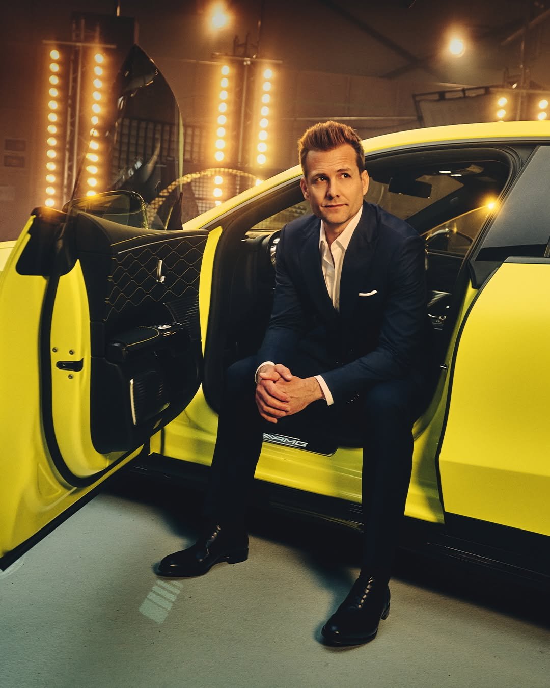 A confident man in a dark suit and white shirt sits on the sill of a bright yellow luxury car. His hands are clasped, and he looks up to the right. The background features rows of glowing lights. 'AMG' is visible on the car's interior.
