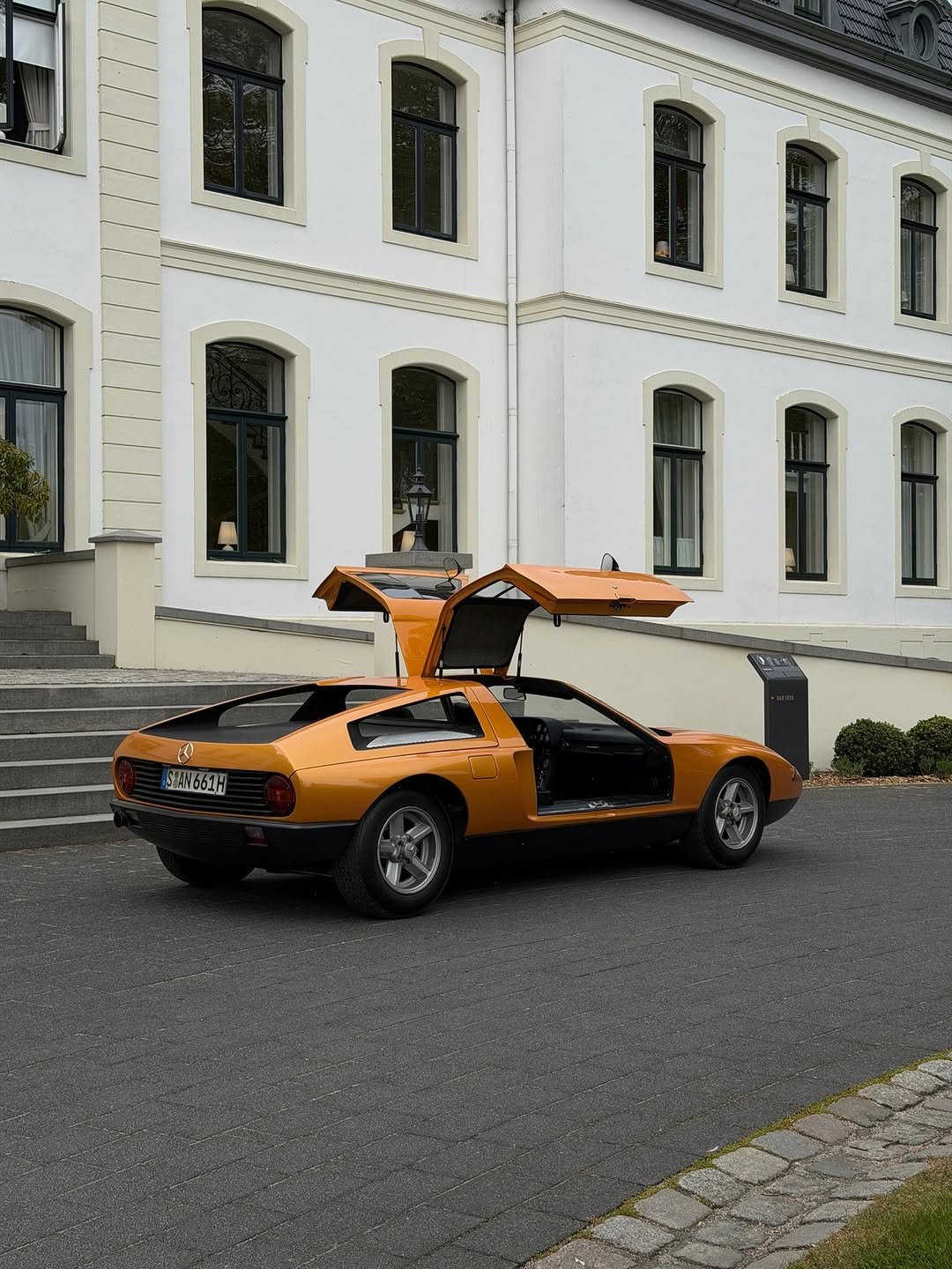 Bright orange Mercedes-Benz sports car with open gull-wing doors parked on dark pavement. It's in front of a grand white building with arched windows and light beige trim. License plate: "S AN 661H". A black sign nearby reads "BAR 1896".