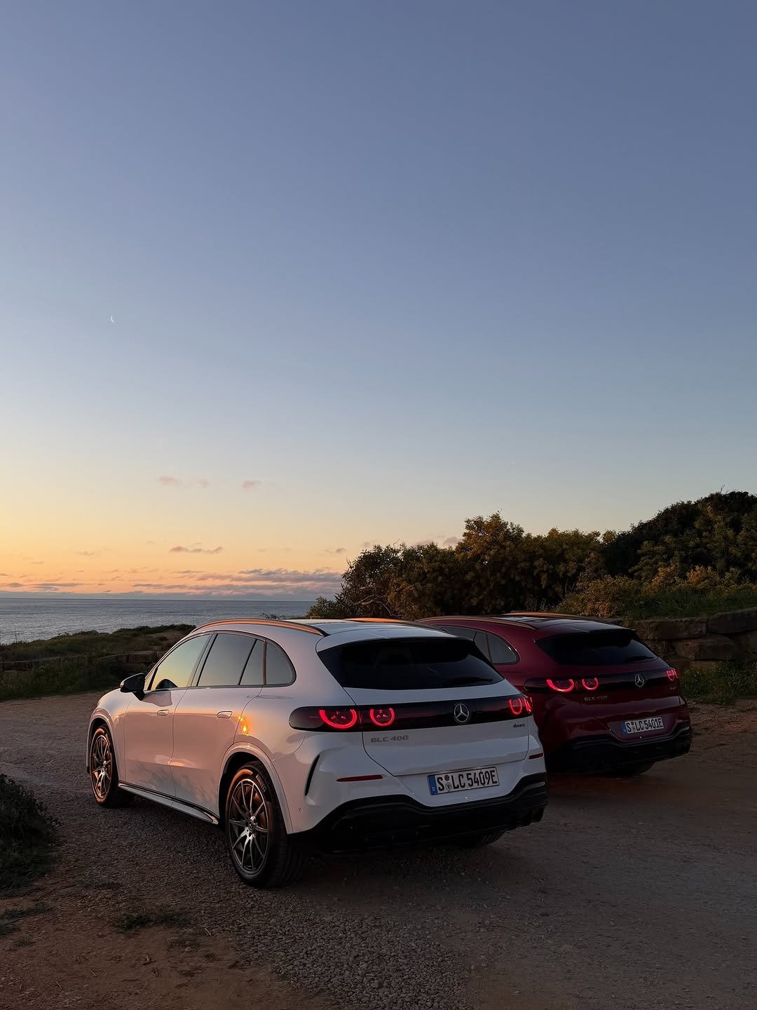 Two Mercedes-Benz GLC 400e 4MATIC SUVs, a white one (S LC 5409E) and a red one (S LC 5401E), are parked on a gravel path by the ocean. The sunset sky fades from orange to blue with a visible crescent moon. Both vehicles have their red taillights illuminated.