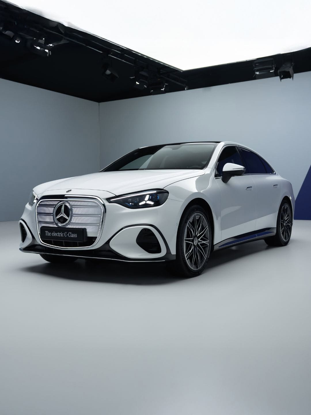 A white Mercedes-Benz electric C-Class sedan is displayed in a brightly lit studio. The car faces slightly left, showcasing its sleek design, prominent grille with the Mercedes logo, and illuminated headlights. 'The electric C-Class' text appears below the logo.