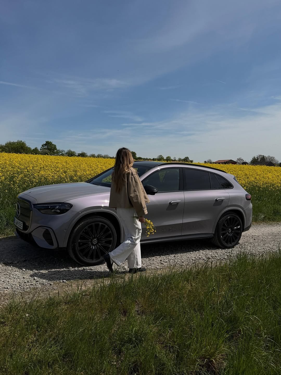 Woman with long hair walks past a silver Mercedes-Benz GLC Coupe on a gravel path, holding yellow flowers. She wears a tan jacket & white pants. A vibrant yellow field is behind the car under a blue sky. License plate: BB ON 855E.