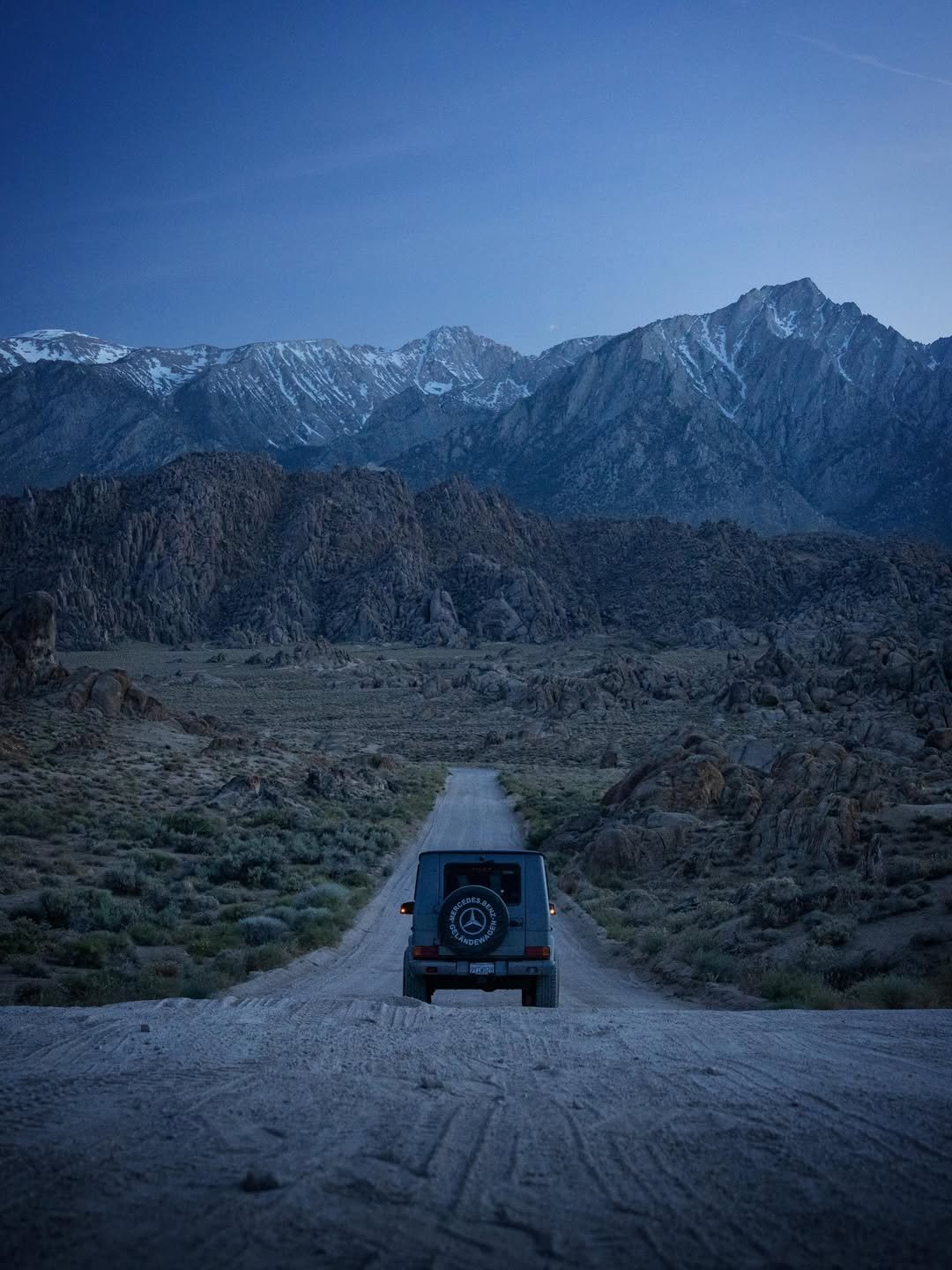 A dark SUV drives away on a dirt road through a rocky, desert-like landscape. In the distance, large snow-capped mountains rise under a deep blue twilight sky. The SUV's spare tire cover has 'Mercedes-Benz Geländewagen' text.