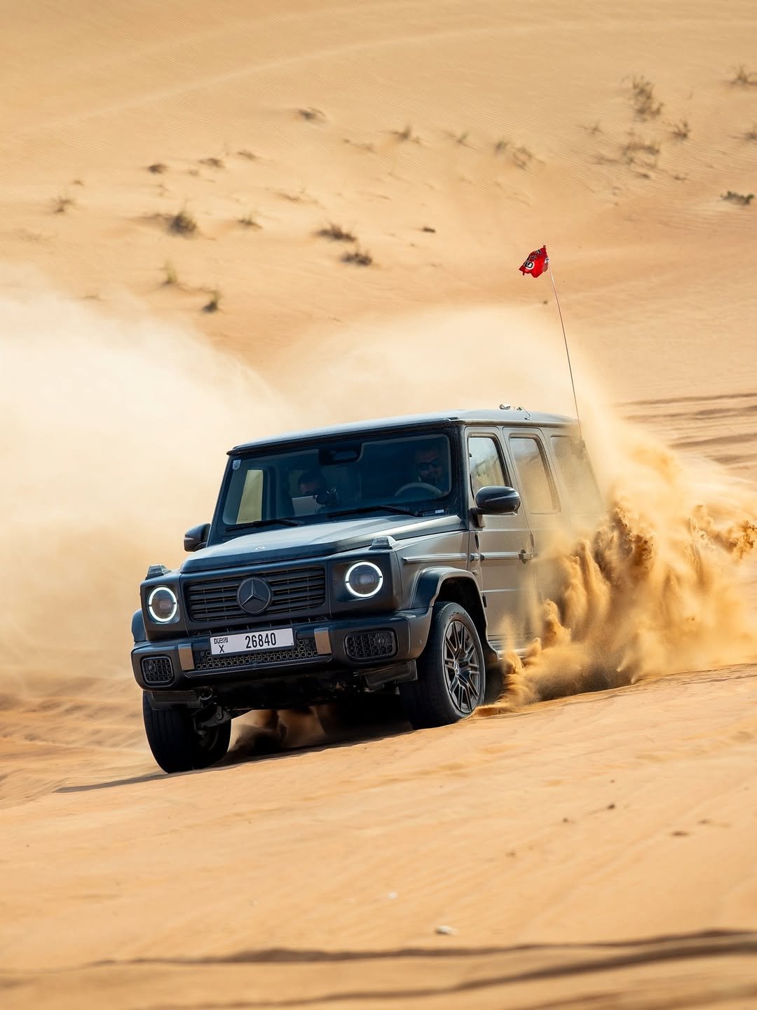 A powerful dark gray Mercedes-Benz G-Wagon blasts through a golden desert dune, kicking up a large plume of sand. Its circular headlights are on, and a red flag flies from its rear. DUBAI X 26840 plate visible, with two people inside.