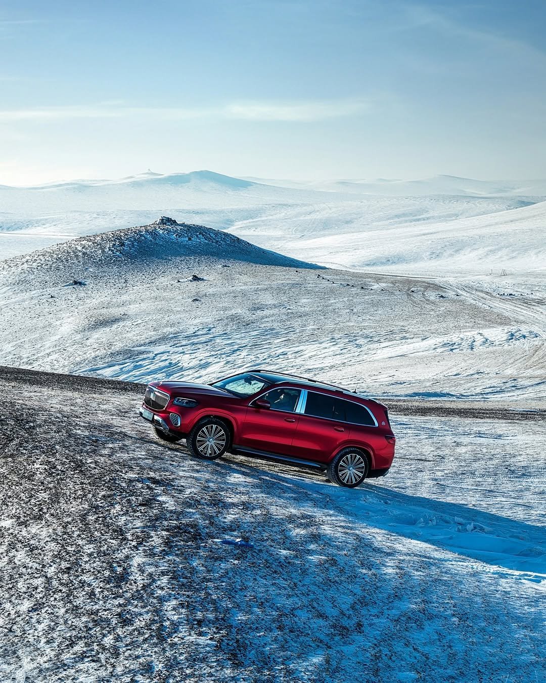 A red SUV, possibly a Mercedes-Maybach GLS, is parked on a snowy, hilly landscape under a clear blue sky. The vehicle is positioned on a slight incline, with snow-covered hills and sparse vegetation visible in the background.