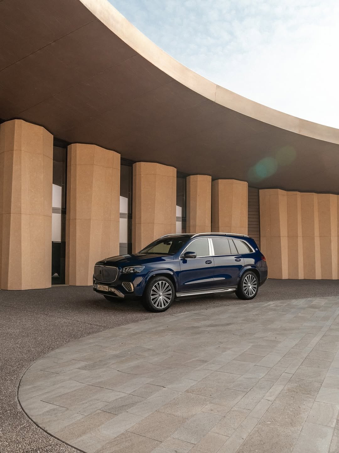 A dark blue Mercedes-Maybach GLS, with plate "S.PX 7894", sits on a paved area in front of a modern building featuring beige columns and an overhanging curved roof. The sky is visible in the background.