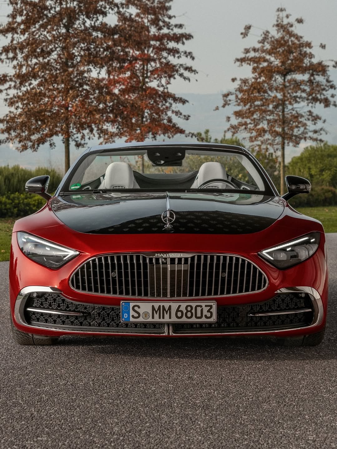 Front view of a red and black Maybach convertible on an asphalt road. The car has a chrome grill with 'MAYBACH' text@1 across the top and a license plate that reads 'S MM 6803' text@2. Two autumn trees are behind it.