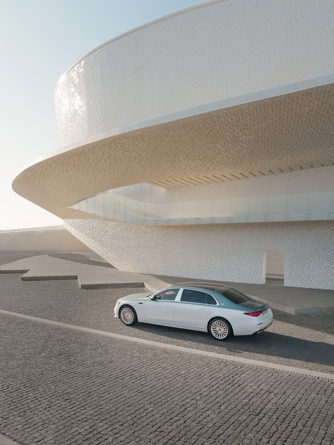 A white and light blue two-tone luxury Mercedes-Maybach S-Class with chrome wheels is parked on dark cobblestones beside a curving, modern building with a textured white facade. Sunlight reflects off the building. License plate reads 'S=MP 2280'.